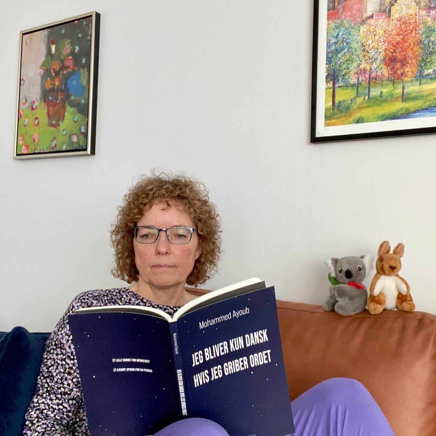 Kvinde sidder iført lilla hyggetøj i cognacfarvet sofa med en digtsamling